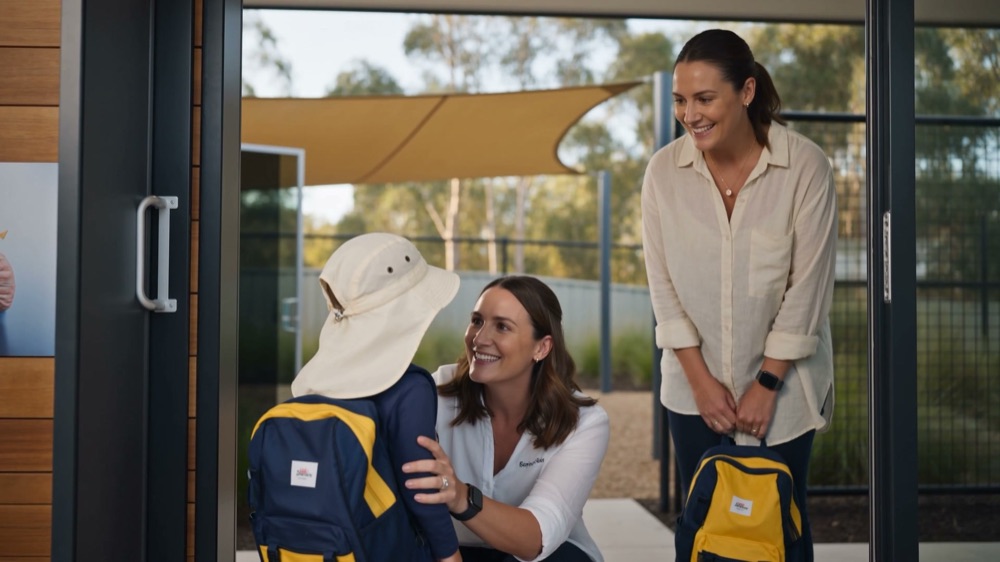 An educator crouching to child height at the front door of the centre to take a backpack, with a parent standing behind