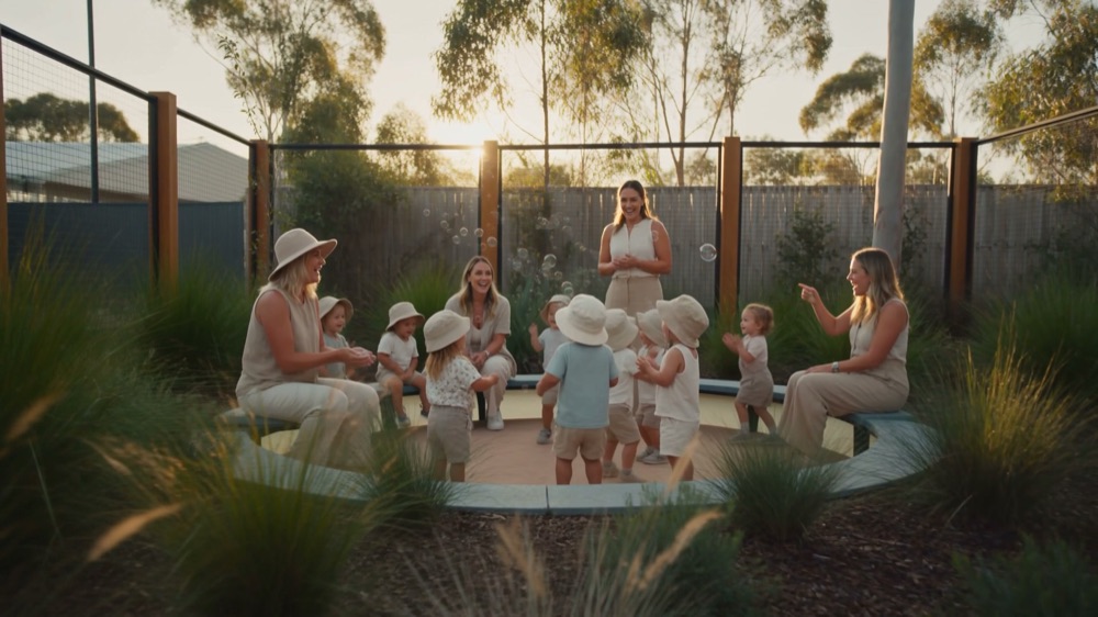 Children and educators sitting in a circle on the grass in late afternoon light with native planting behind them