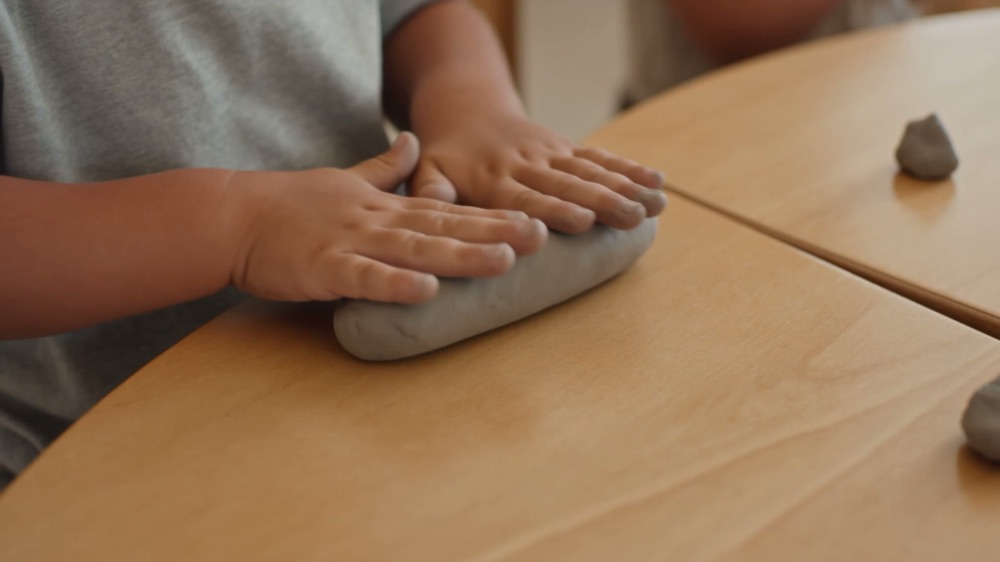 A small child pressing both hands flat into a slab of grey clay on a plywood table